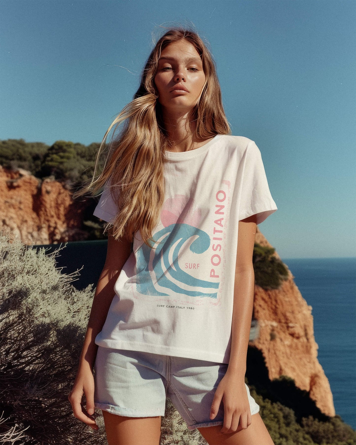 Woman wearing a 'POSITANO' t-shirt with a graphic design, standing on a cliff overlooking the ocean.