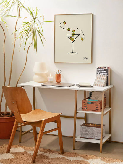 A mid-century modern writing desk with a white top and gold metal frame, styled in a warm room with a wooden chair, a potted plant, and a framed art print on the wall.